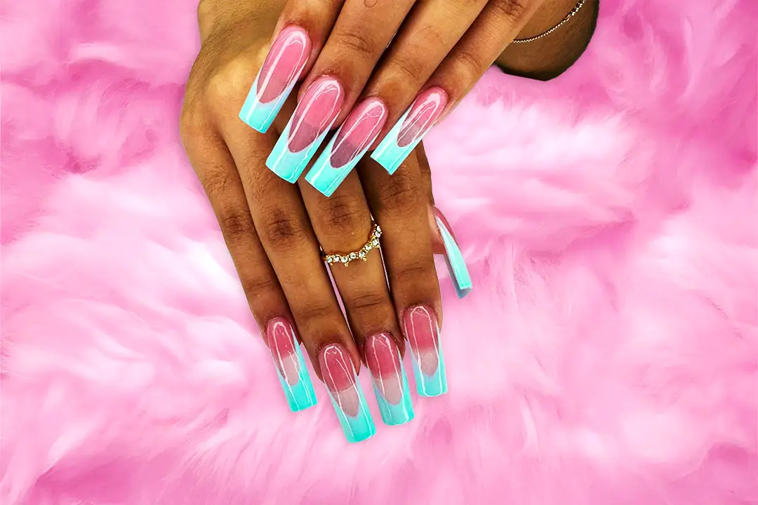 A close-up view of a hand with long, manicured nails painted in blue, pink, and turquoise, adorned with a ring on a soft pink fur background.