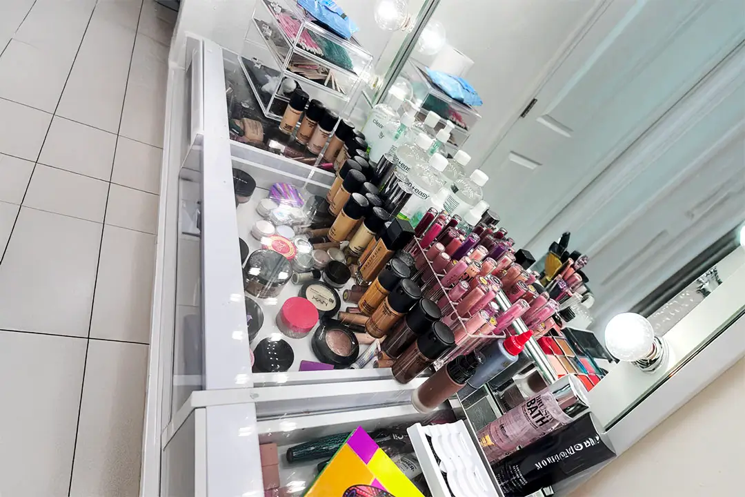 Well-organized makeup counter with white shelves, mirror, lipsticks, eyeshadows, foundation, and magazines arranged on a tiled floor.