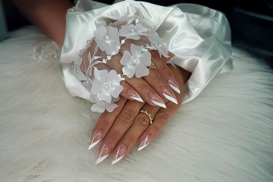A persons hand with long white nails, silver rings, and a black and white tattoo on the thumb, holding a bottle and a mirror.