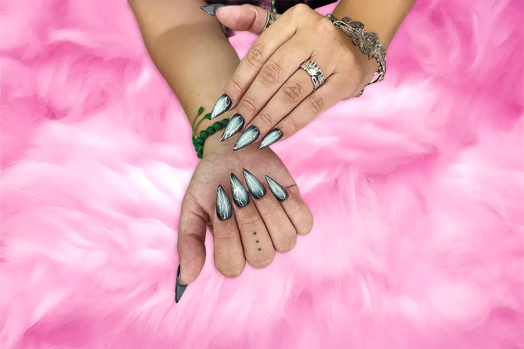 A hand with long, metallic silver nails, adorned with green and black polish, rests on soft pink fur, wearing a silver bracelet and ring.