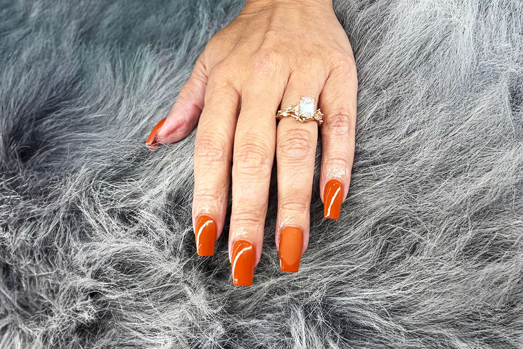 A hand with vibrant orange nail polish and a gold diamond-shaped ring rests on a soft gray fur surface.