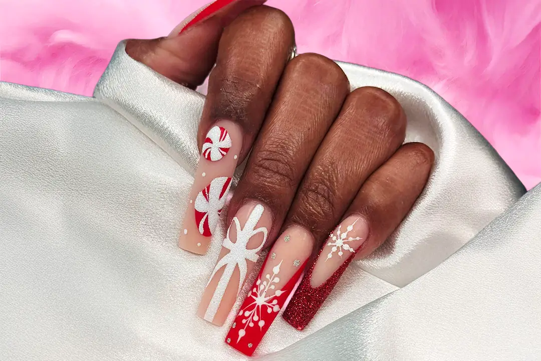 A hand with festive red and white nail art featuring snowflakes, stars, and bows on a silver surface against a soft pink background.