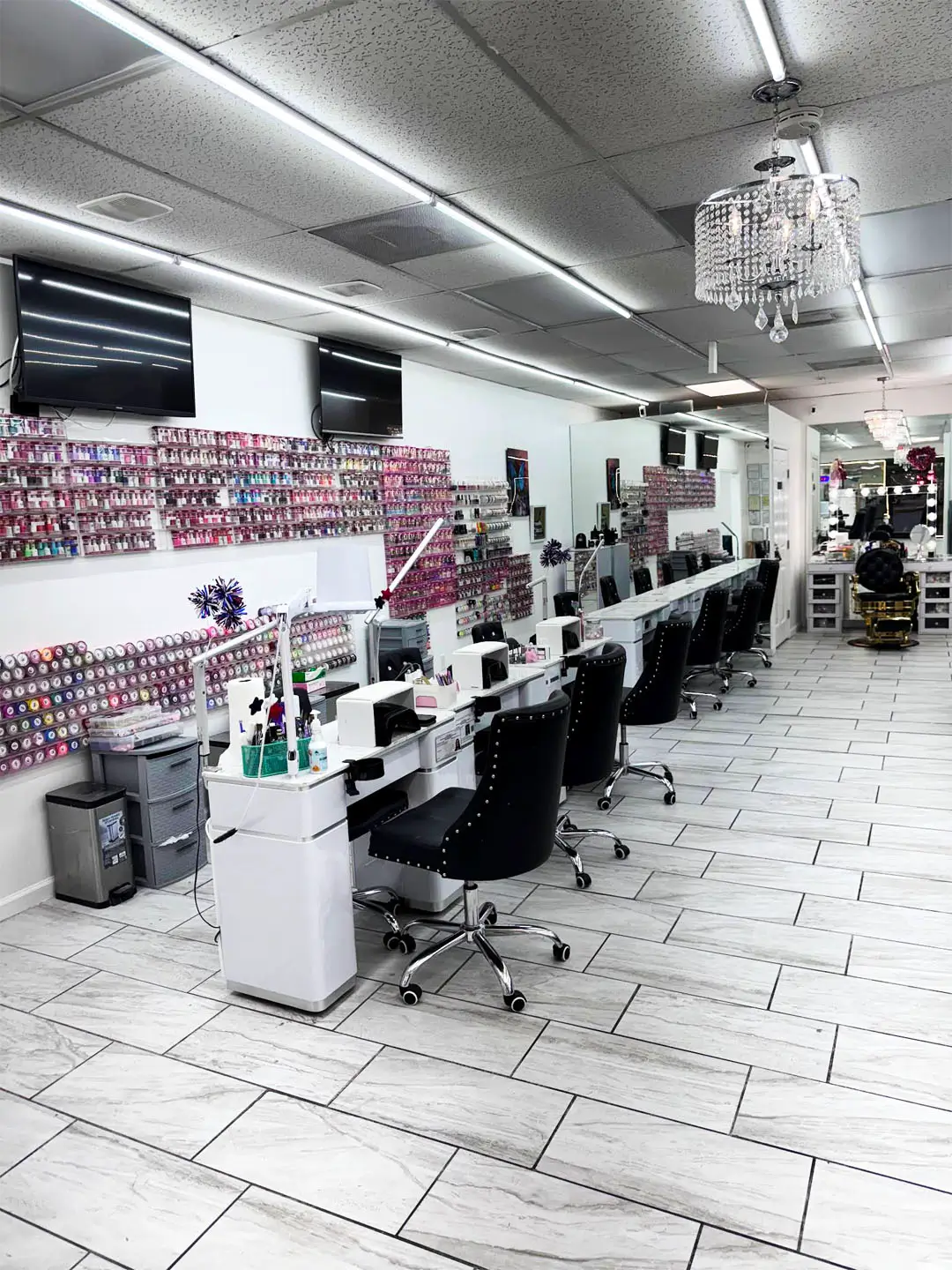 Well-lit salon with white tiles, chandelier, lamps, hairdryers, hairbrushes, and diverse hair products displayed on the walls.