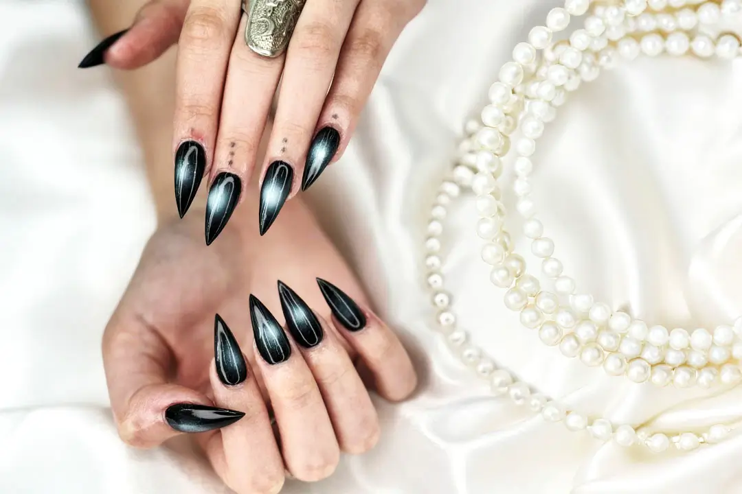 A persons hands with black nail polish and a gold ring rest on white fabric, near a pearl necklace.